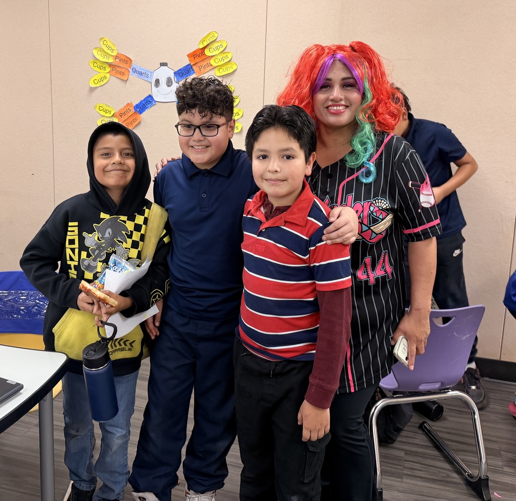 A medium indoor photograph shows four people standing together near a table. On the left, a smiling young boy wears a black Sonic the Hedgehog hoodie, holds a slice of pizza on paper, and carries a blue water bottle. Next to him is another smiling boy with glasses wearing a navy polo. A third, younger boy, smiles from the front-right wearing a red and navy striped shirt. On the far right stands a smiling PTSO volunteer with bright red and teal hair, wearing a striped baseball jersey and placing an arm around one of the boys. In the background is a beige wall with a colorful classroom measurement-chart craft.