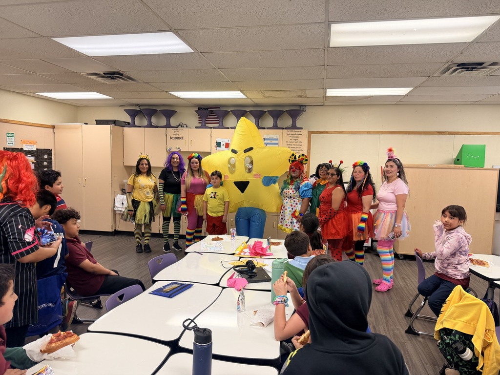 A wide-angle indoor photograph captures a busy elementary school classroom. In the background, a large yellow inflatable star mascot is posing with several of the costumed PTSO volunteers seen in previous images. In the foreground, students are sitting at white tables, talking, and eating slices of pizza. One student is wearing a blue hooded sweatshirt. The classroom is filled with desks, chairs, and shelves, under fluorescent lights and a drop ceiling.