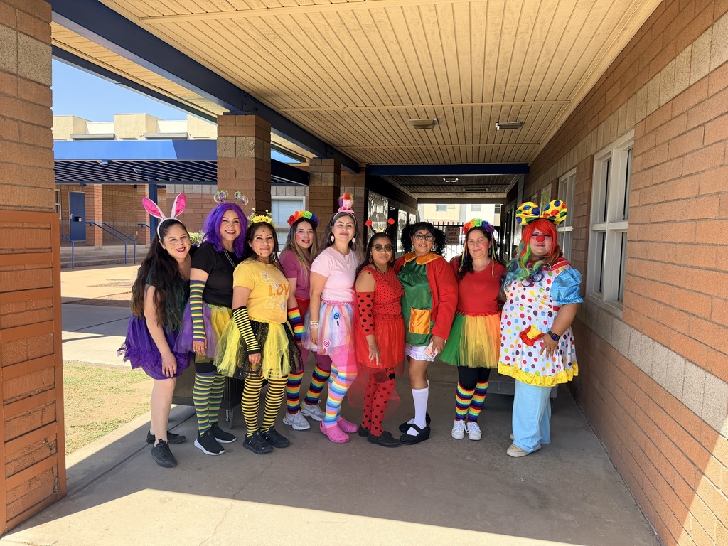 A wide, sunlit photograph taken outdoors on a school sidewalk shows nine female PTSO volunteers lined up in various colorful costumes. From left to right, their costumes include a purple rabbit, a purple-haired bee, a yellow bee with a tutu, a pink fairy, a ladybug, an M&M, and a clown in a polka-dot dress. They are smiling and posing under a shaded brick walkway next to a modern school building with blue awnings.