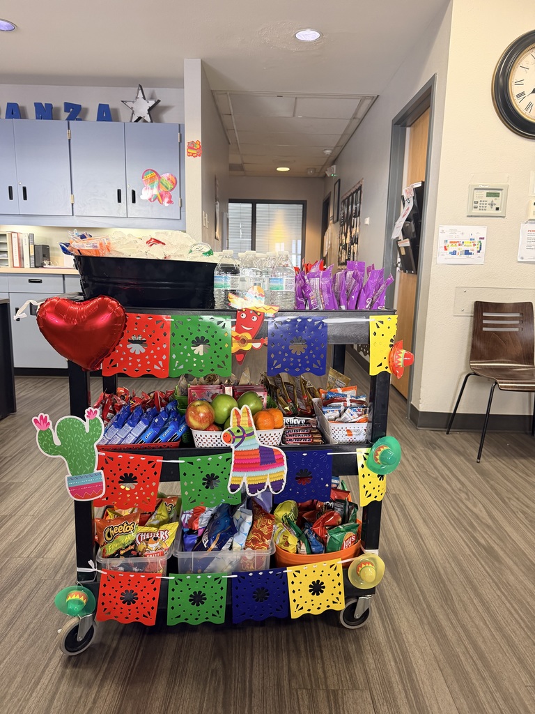 A three-tier black rolling utility cart heavily decorated for a "Fiesta" theme. The cart is adorned with colorful papel picado banners in red, green, blue, and yellow. Decorations include a red heart balloon, a cardboard cactus, a colorful piñata donkey, and several small sombreros.  Top Tier: A large black tub filled with ice and bottled water, alongside purple bags of snacks.  Middle Tier: Red baskets filled with Oreo packs, fresh green apples, oranges, and various chocolate bars (Hershey’s, Snickers, KitKats).  Bottom Tier