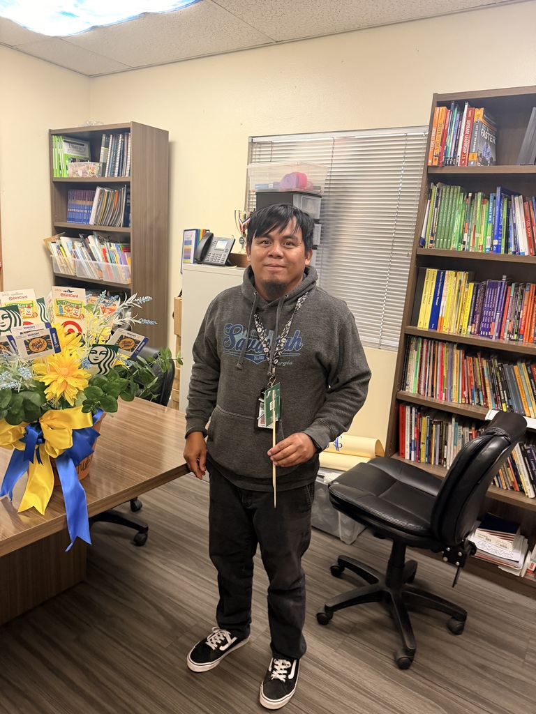 A man wearing a dark grey "Savannah" brand hoodie and black pants stands in the office. He has a calm smile and is holding a wooden stick that was used for the gift card raffle. The colorful gift card bouquet with yellow flowers and blue/yellow ribbons is positioned to his left on the wooden desk.
