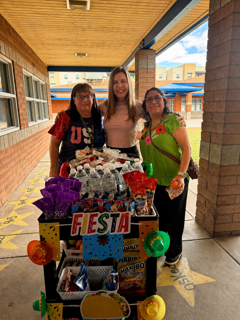 Three women stand together outdoors under a school walkway, smiling warmly behind the fully loaded Treat Trolley. The woman in the center has her arms around her colleagues, celebrating the day’s successful snack deliveries.