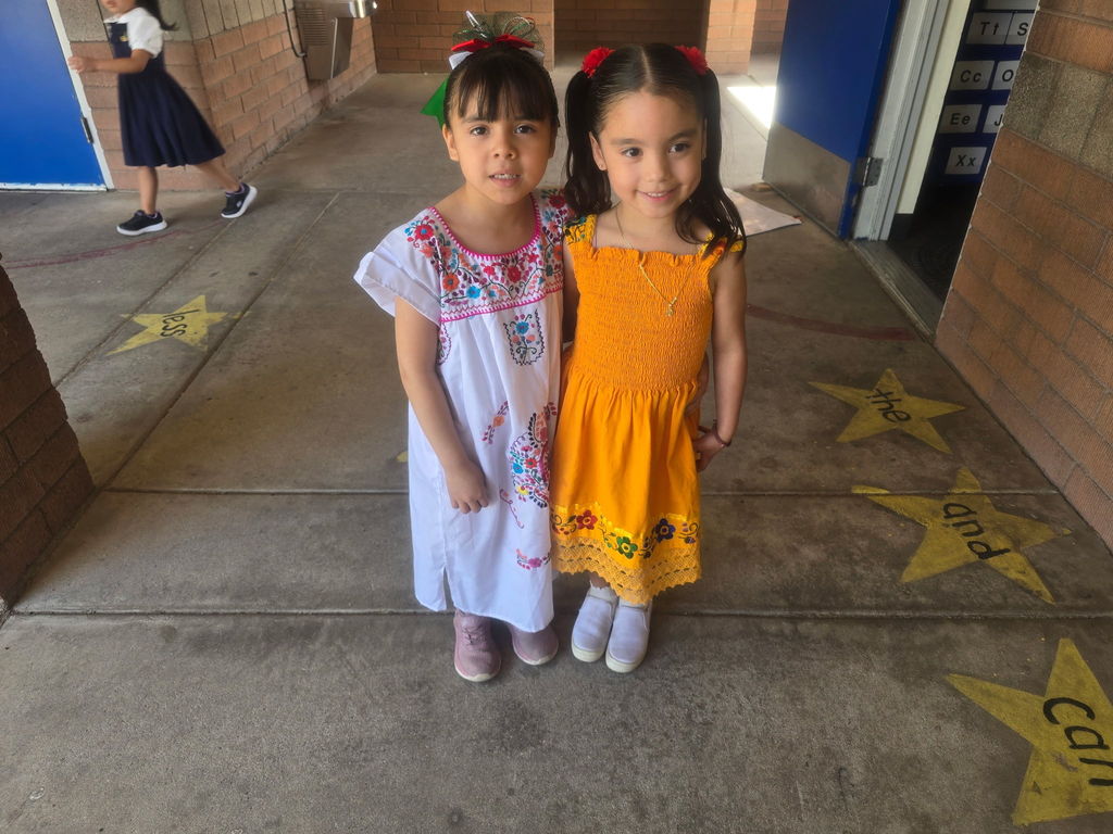 Two young girls smiling together. One wears a white "huipil" style dress with multicolored floral embroidery, and the other wears a bright orange smocked dress with floral details at the hem.