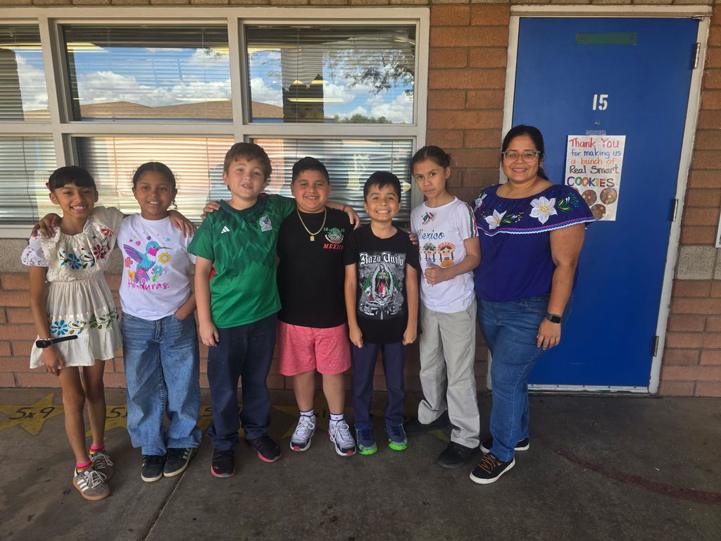 A group of six students and one female adult posing outside a classroom. Many are wearing white embroidered tops or soccer jerseys representing Mexico and Honduras.