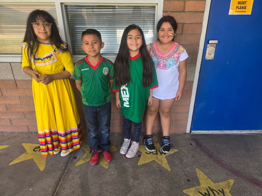 Four students standing in a row. They are wearing a yellow folklorico-style dress, a green Mexico soccer jersey, and a white embroidered blouse.