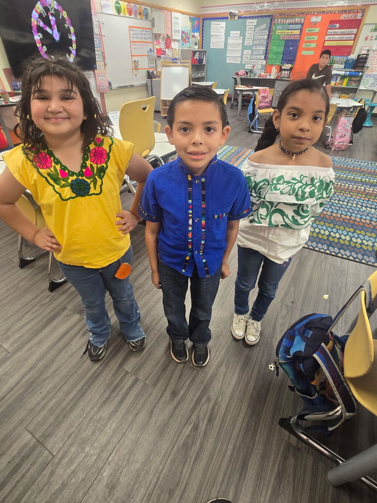 Three elementary students standing in a classroom. A girl wears a yellow blouse with pink floral embroidery, a boy wears a bright blue button-down shirt with traditional patterned trim, and a girl wears a white off-the-shoulder blouse with green floral patterns.