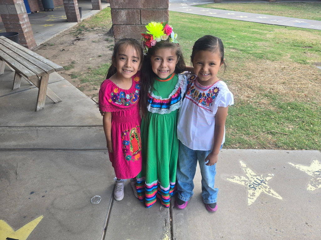 Three young girls standing outside on a sidewalk with star decals. They are wearing traditional Mexican dresses in vibrant shades of pink, green, and white, featuring intricate floral embroidery and ribbon tiers.
