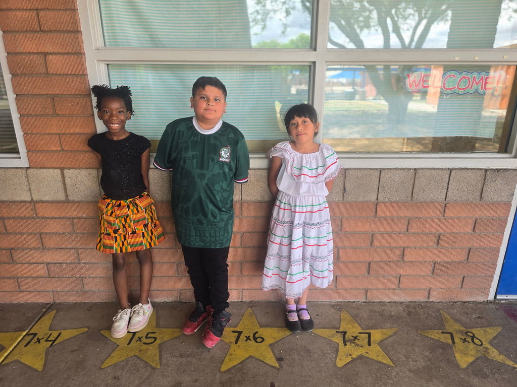 Three students standing over star decals on the pavement. One girl wears a black top and a vibrant orange and yellow patterned Kente-style skirt, a boy wears a dark green patterned jersey, and a girl wears a white tiered dress with red and green trim.