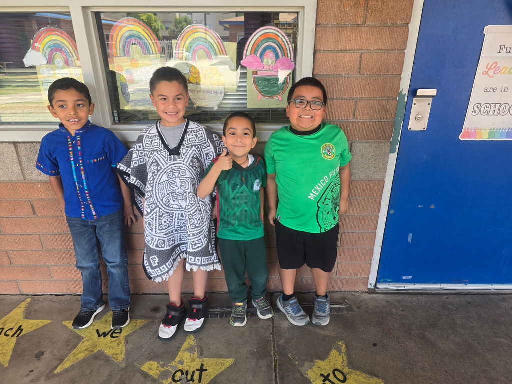 Four boys standing against a wall. Their outfits include a blue traditional shirt, a grey and white patterned poncho (jorongo), a green Mexico soccer jersey, and a bright green t-shirt.