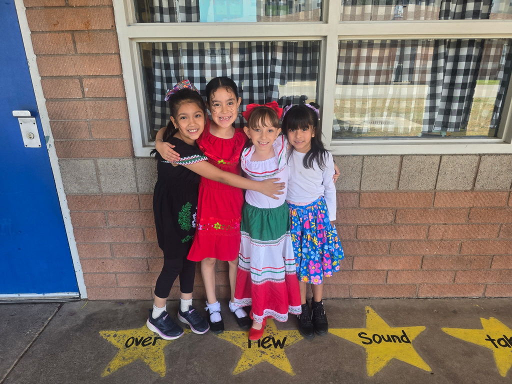 Four girls posing together in a hug. They are dressed in various cultural attires, including a red ruffled dress and a white blouse with a green, white, and red ribbon-tier skirt.