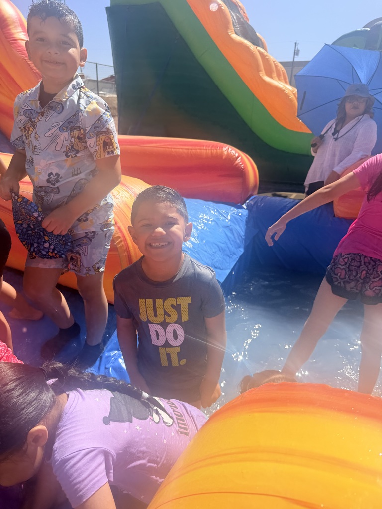 A close-up shot during a school Water Day event. In the center, a young boy with wet hair wears a dark grey shirt that reads "JUST DO IT" in yellow and white text, smiling brightly at the camera. He is standing in the shallow water pool of an orange and blue inflatable water slide. Other children are splashing nearby, including a boy on the left in a blue patterned shirt and a girl in a purple shirt in the foreground. In the background, a person holds a blue umbrella under a clear, bright sky.