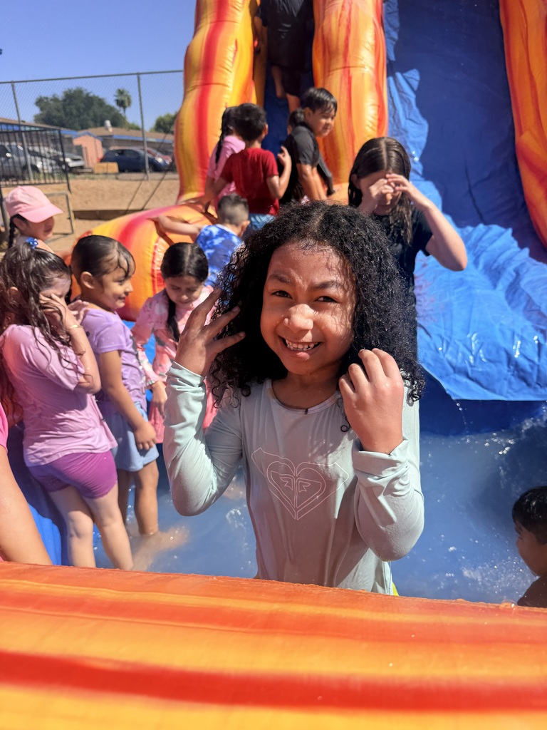 A close-up shot of a young girl with long, wet, curly dark hair smiling brightly at the camera. She is standing in the shallow water of an inflatable pool, with her hands near her face. In the background, out of focus, several other children play and climb on a red and orange inflatable water slide.
