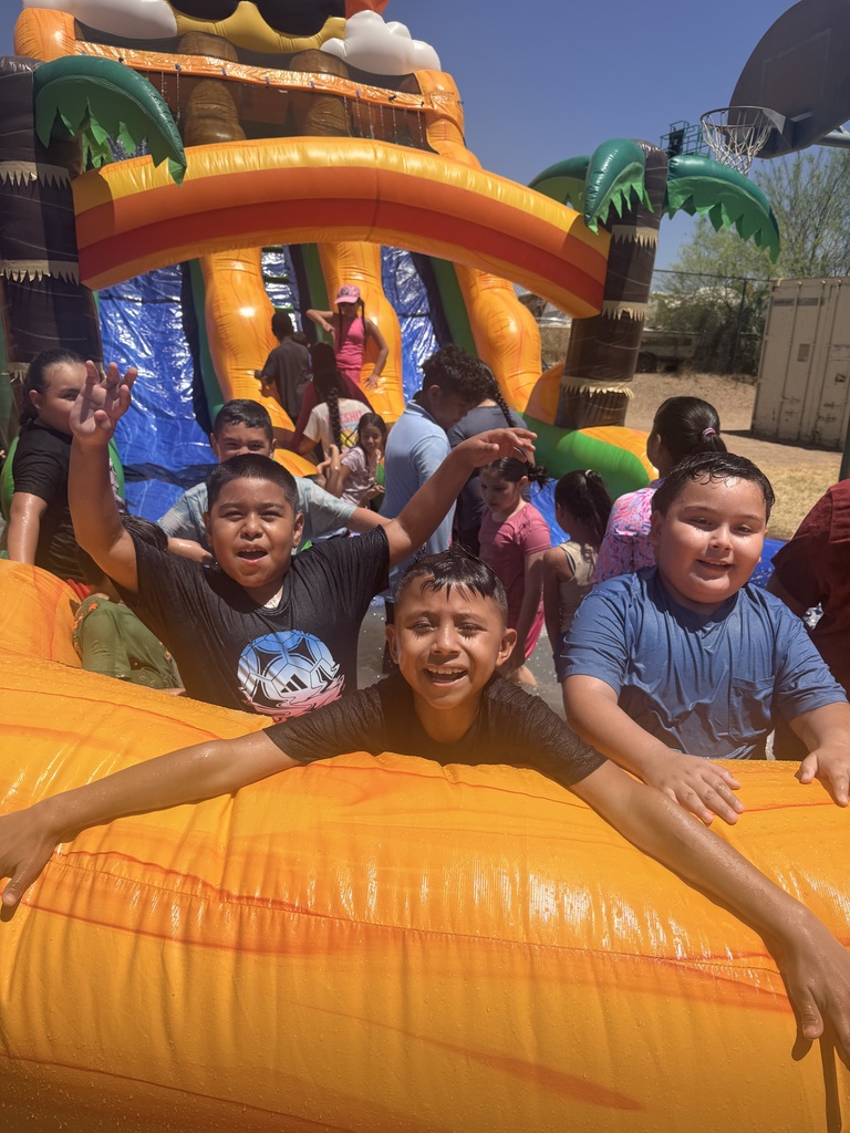Three wet, smiling boys lean over the front orange barrier of an inflatable water slide pool, looking excited. The boy on the left has his arms raised high in mid-splash. Behind them, the large tropical-themed inflatable water slide—complete with palm trees and a smiling sun wearing sunglasses—is filled with other children playing on a clear, sunny day.