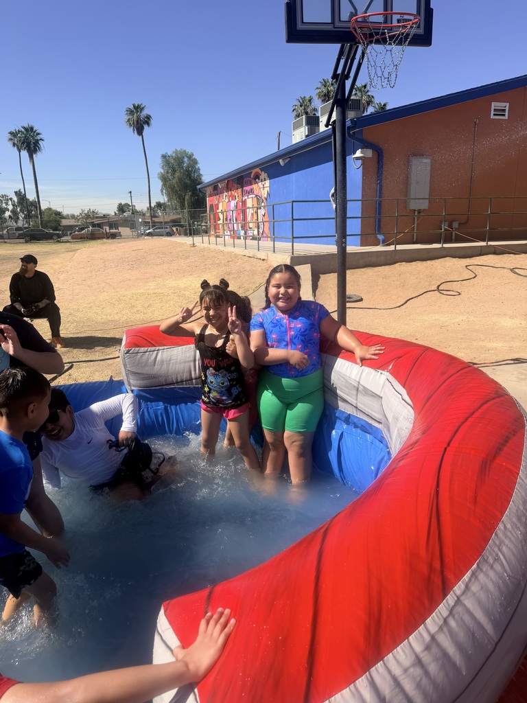 Two young girls stand inside a red and grey inflatable splash pool on a sunny day, smiling and flashing peace signs at the camera. One girl wears a black swimsuit and the other wears a blue patterned rash guard with green swim shorts. A basketball hoop stands directly behind the pool. In the background, a blue and orange school building features a colorful painted mural under a clear blue sky.
