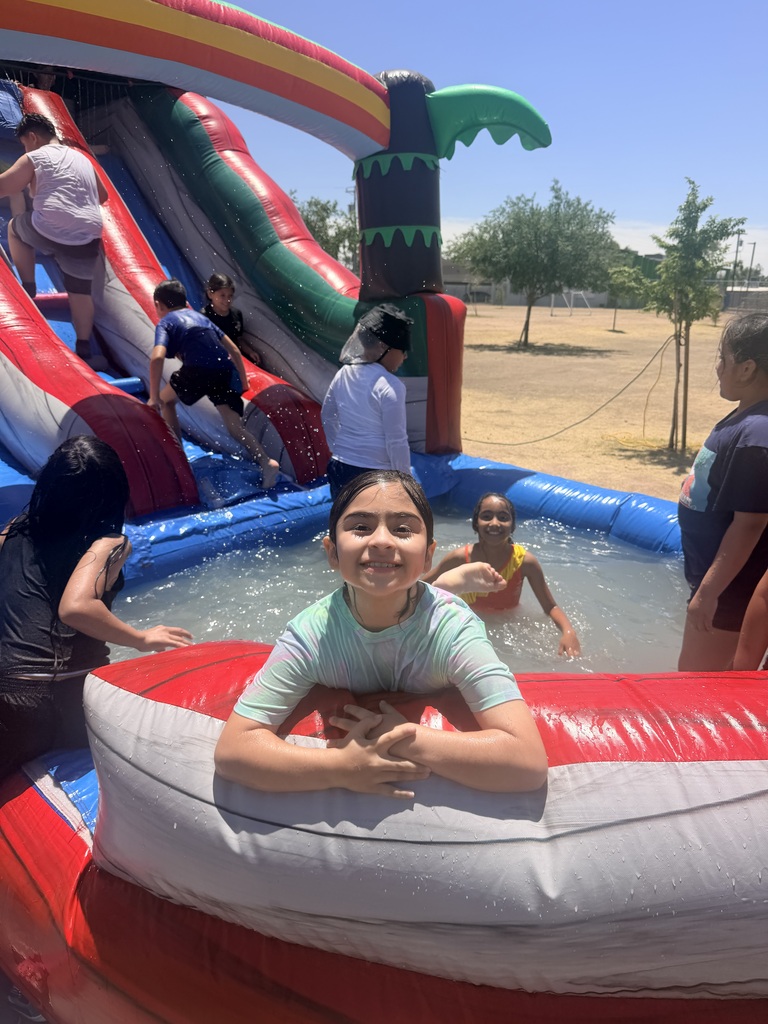 A close-up of a young girl with dark hair leaning against the grey and red rim of an inflatable water slide pool, smiling warmly with her hands clasped. In the pool behind her, another girl in an orange swimsuit smiles at the camera. To the left, children climb up the steps of the red and blue water slide. The background shows an open, sandy school field with a few young trees under a bright blue sky.