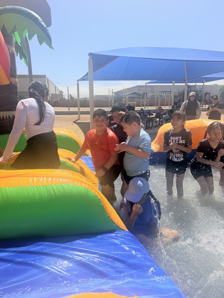Several children play and splash at the base of a green and blue inflatable water slide. Two boys, one in a red shirt and one in a light blue shirt, stand near the slide's exit ramp. In the background, a large blue canopy shade structure covers a playground area where a few adults stand and supervise the event on a clear, sunny day.