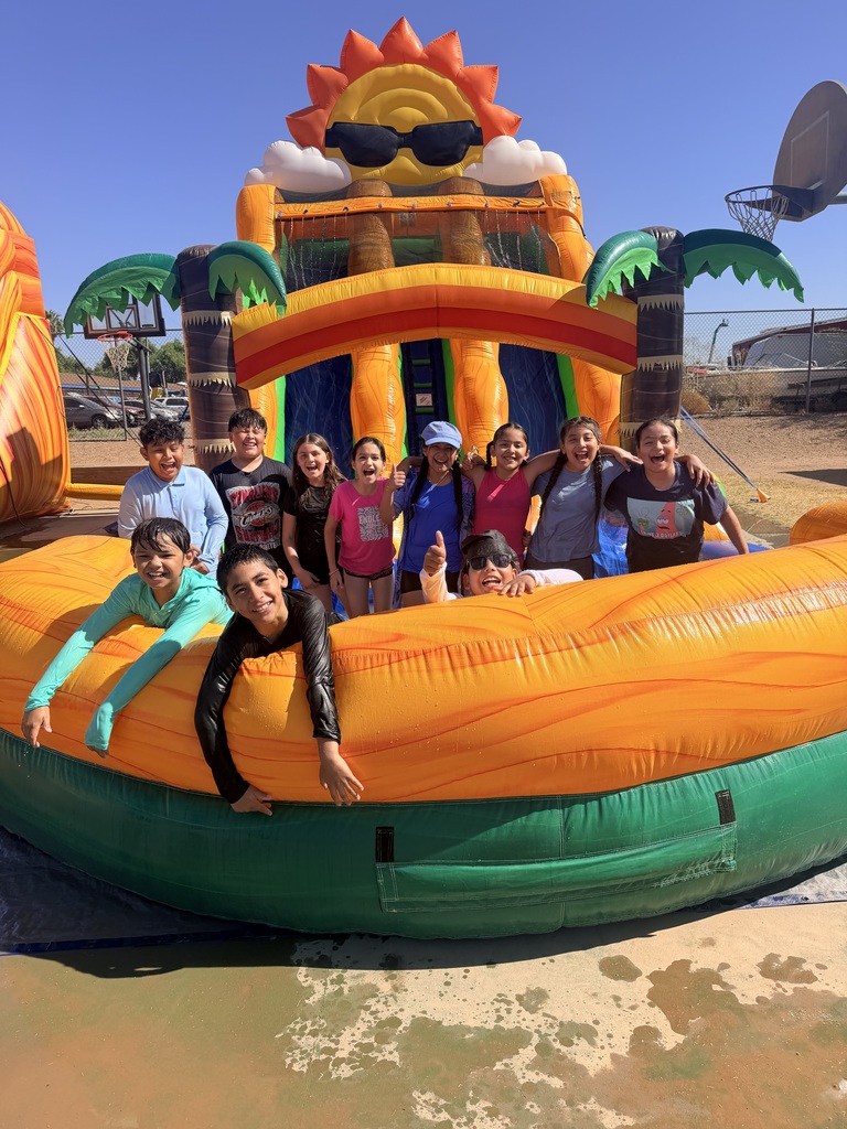 A wide group shot of about ten elementary students packed together, laughing and posing inside the front splash pool of a giant inflatable water slide. The slide features an archway with inflatable palm trees and a large, smiling cartoon sun wearing sunglasses at the top. The children are soaking wet, giving thumbs-up signs and smiling widely on a bright, sunny day.