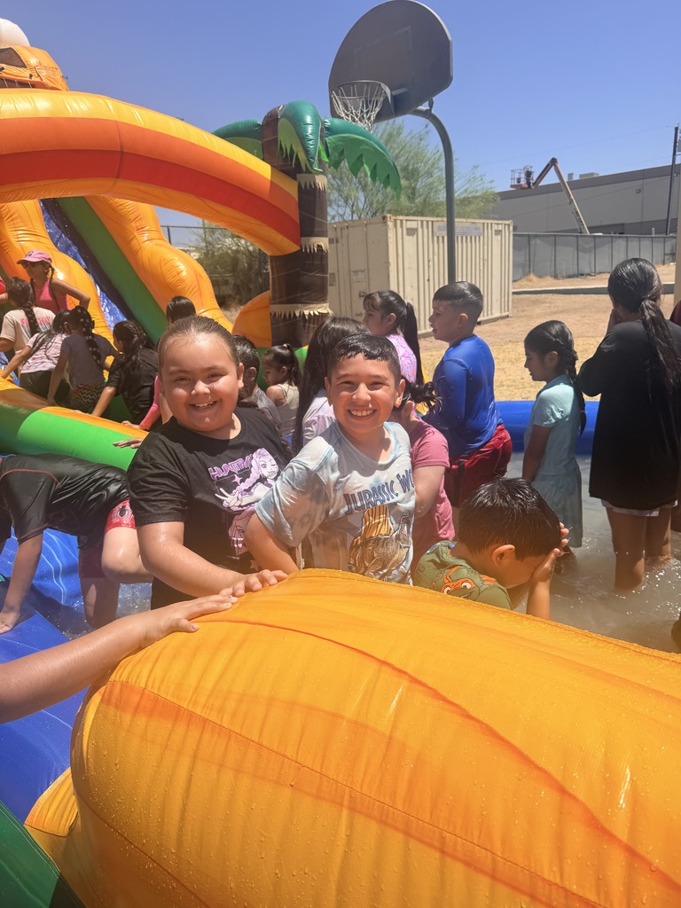A boy in a wet white graphic t-shirt and a girl in a black t-shirt smile happily at the camera from inside a crowded inflatable splash pool. They are surrounded by other children splashing in the water. An orange inflatable slide structure stands to the left, and a basketball hoop and a beige storage container are visible in the background under a bright, clear sk