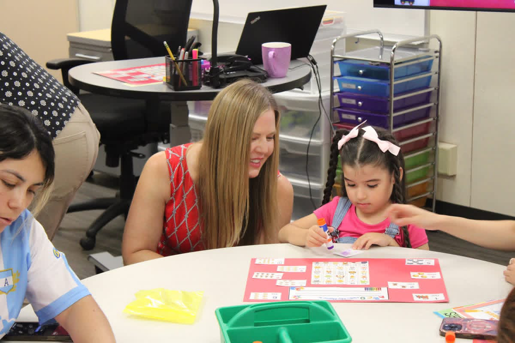 An adult smiles and supports a young student wearing pink and denim overalls as she uses a glue stick to complete a colorful learning activity at a classroom table.