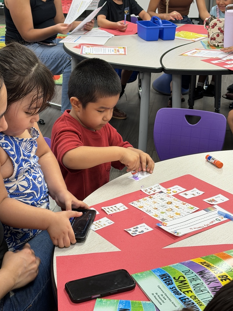 A young student in a red shirt uses a glue stick to complete a math activity on red construction paper. Family members sit nearby, assisting and observing as other participants work around the classroom.