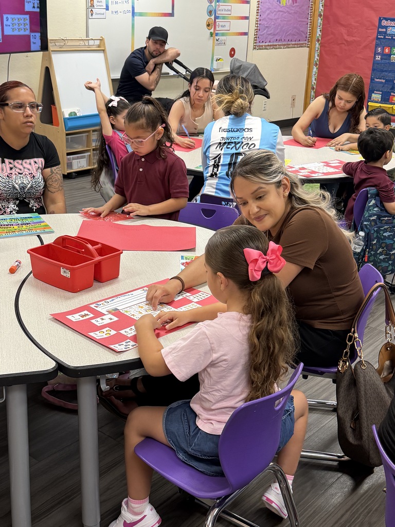 Families and students work together at classroom tables on colorful learning activities. An adult helps a young student complete a number-based project while other families participate in the background.