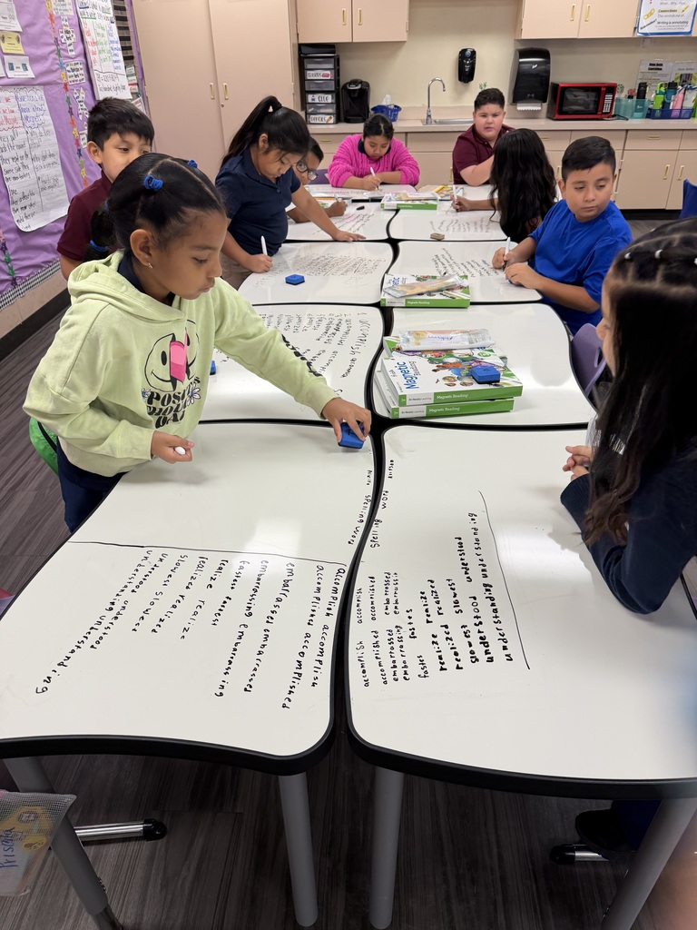 A classroom photo shows a group of third-grade students gathered around several connected whiteboard-top tables. Students are writing words and notes directly on the tables with dry-erase markers, while one student uses an eraser to clear part of a table. Reading materials and classroom supplies are placed in the center. The students are actively working together on a literacy activity in a bright classroom.