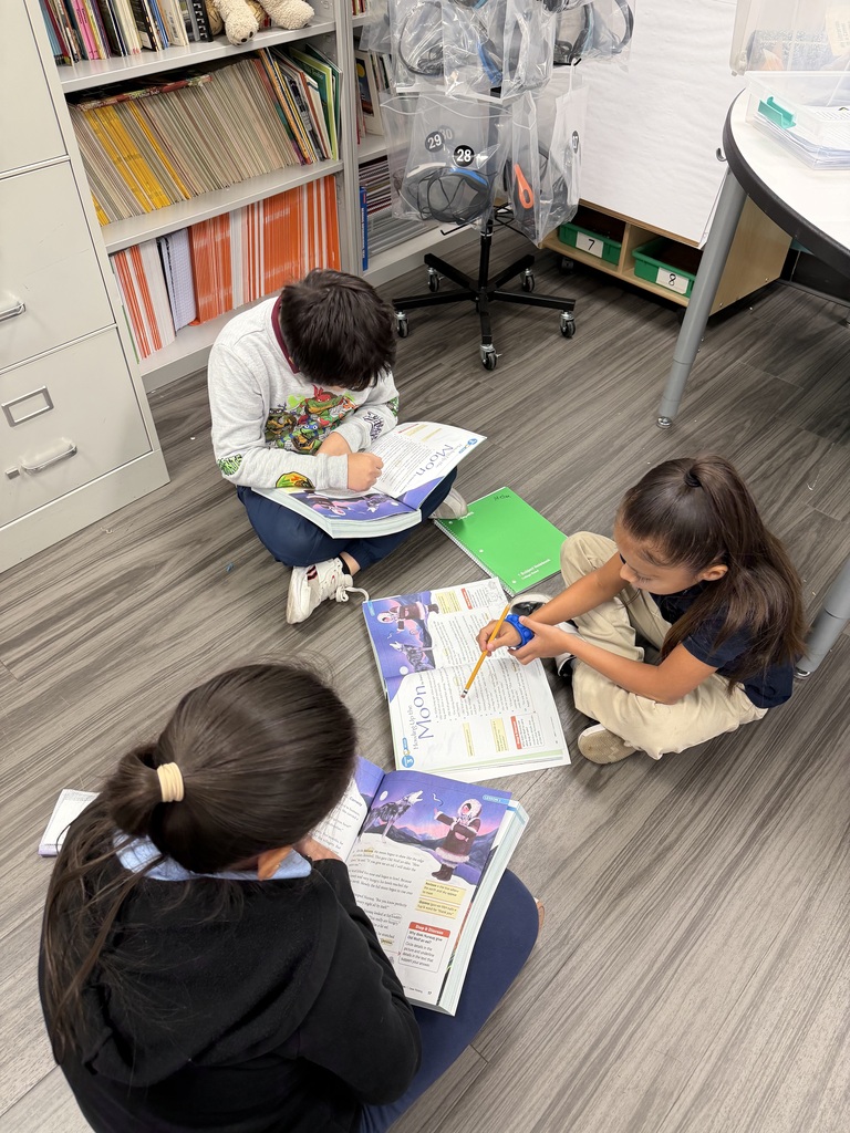 A classroom photo shows three third-grade students seated together on the floor with open reading books. Two students are looking closely at their books while another student points to the text with a pencil and holds a small blue classroom tool. Bookshelves, notebooks, and classroom materials can be seen around them. The students are engaged in a small-group reading and learning activity.