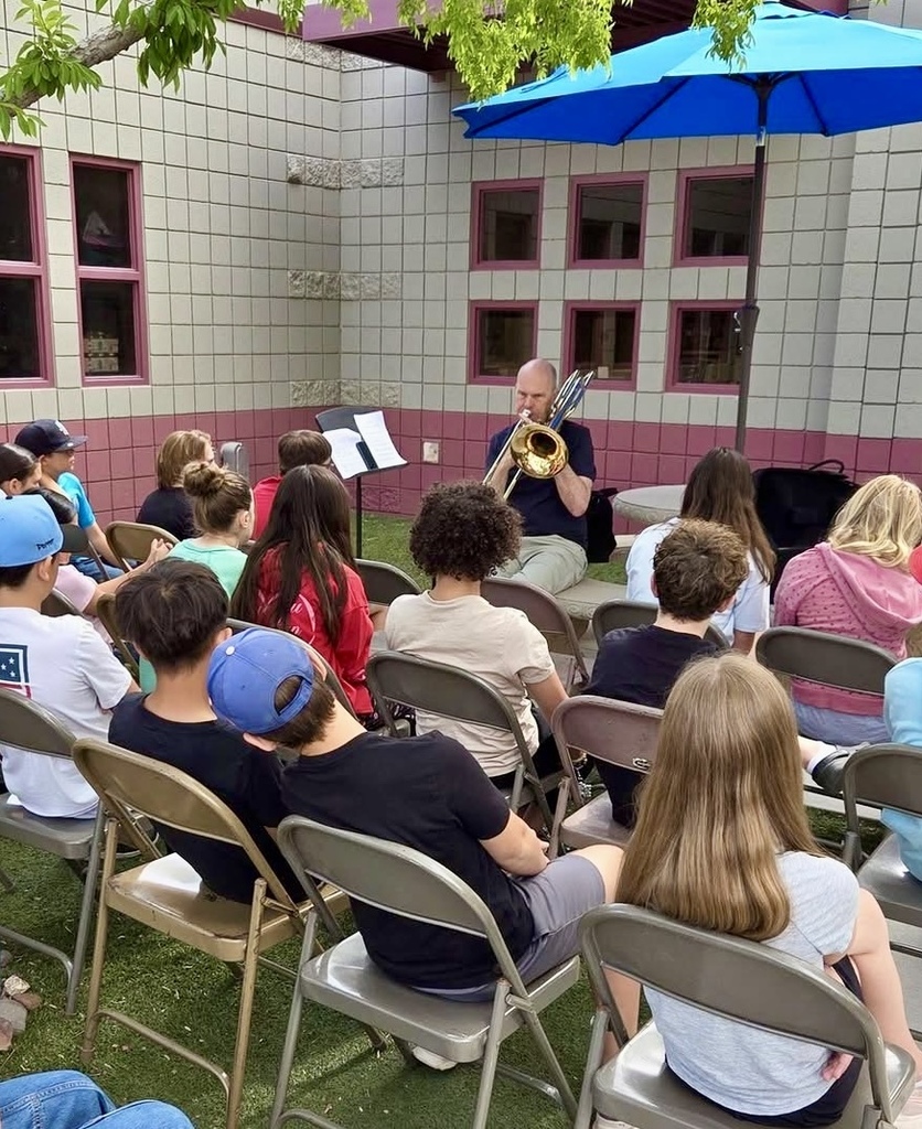 Group of children sit in chairs watching a man play a trumpet under a blue umbrella.