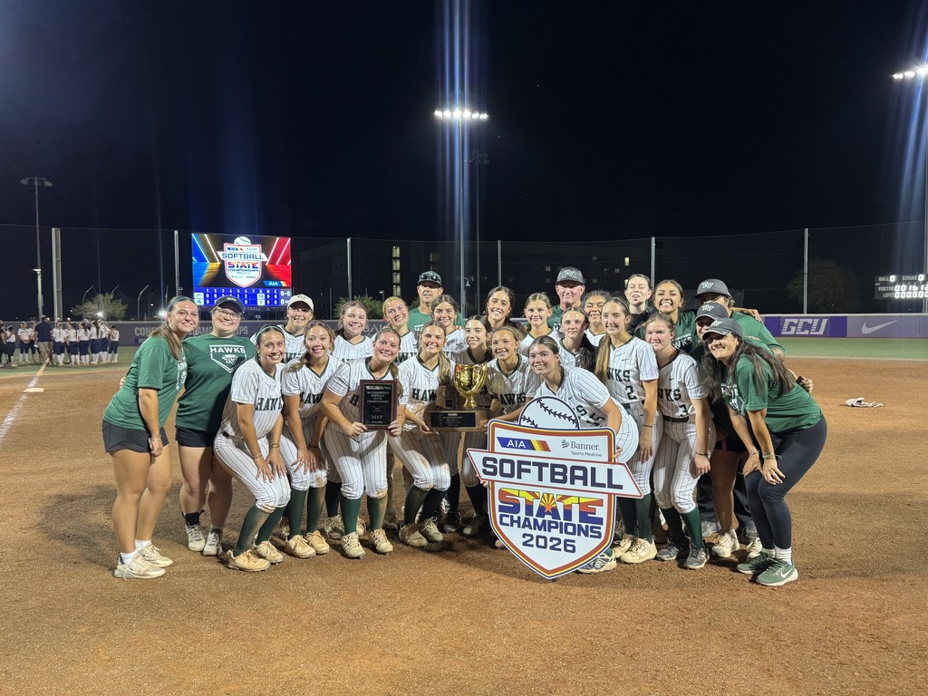 Softball team in green and white uniforms standing in a circle around a trophy on a field.