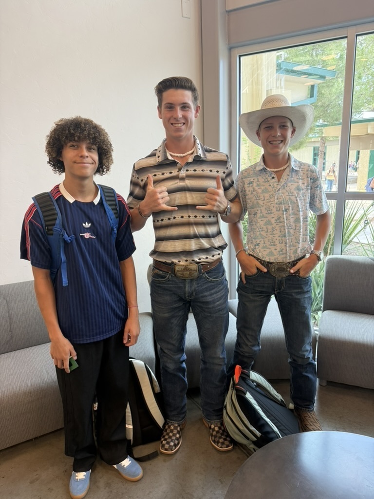 Three individuals stand in a room with backpacks, two giving thumbs-up. One wears a hat, another a necklace, and all wear jeans.