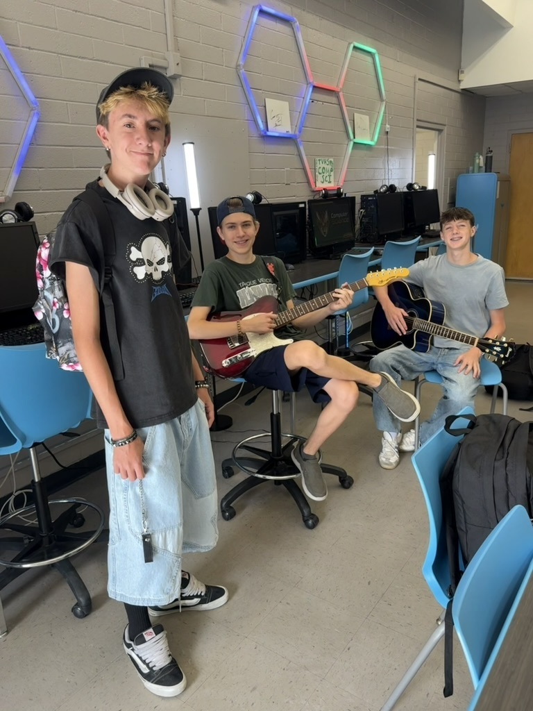 Three young men pose with guitars in a room with hexagonal lights, blue chairs, and monitors.