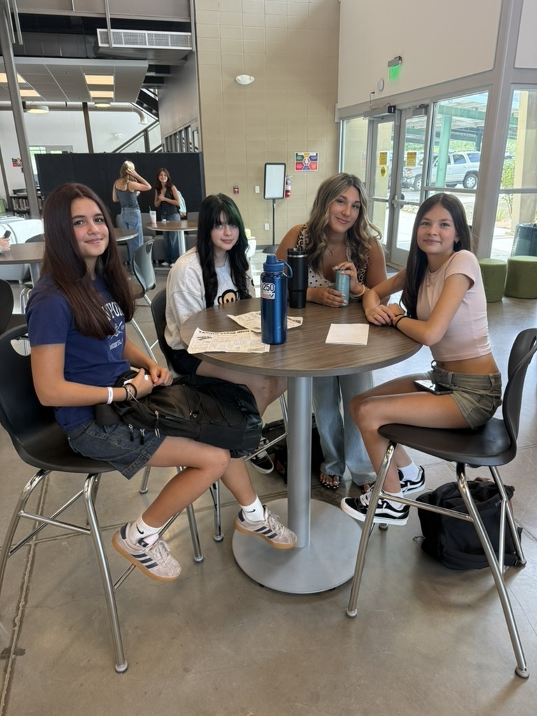 Four women in casual clothes sit at a round table in a cafeteria. Two wear sneakers.