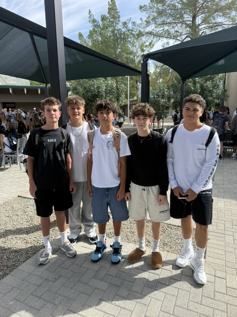 Five young men stand next to each other in an outdoor area with brick floors. They are dressed casually.