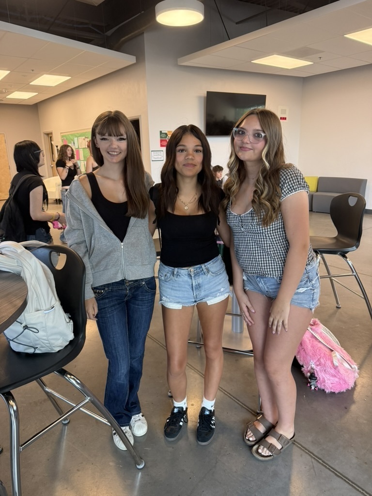 Three women stand in a room, two with arms around each other, and another standing nearby. Chairs, tables, and bags are in the background.