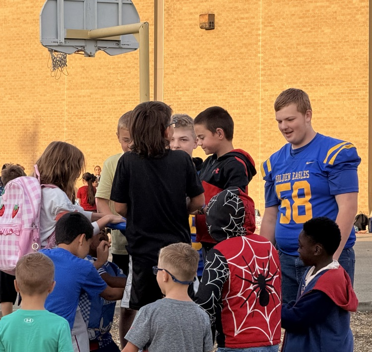 High school football players surrounded by elementary students outside.