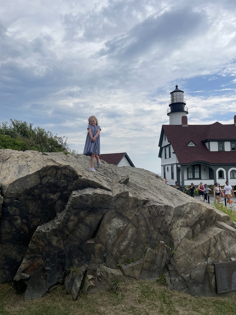Elizabeth is standing in the top of huge rocks with a lighthouse and tourists in the background
