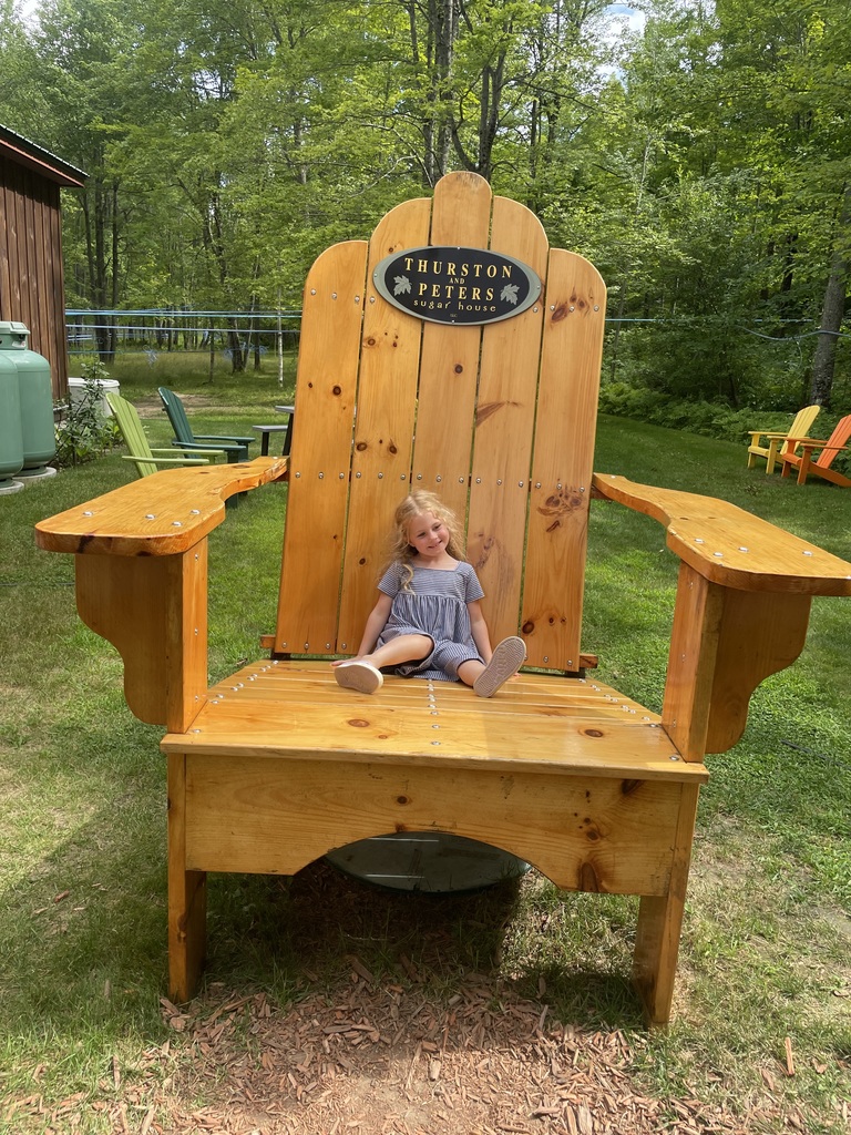 Elizabeth is sitting on a huge over-sized wooden lawn chair smiling with additional lawn chairs on the grass in the background, a sign on the chair reads, "Thurston and Peters Sugar House"