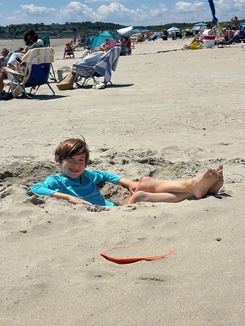 Jensen sits in a hole dug into the sand, wearing a blue shirt and smiling, with other beachgoers in the background