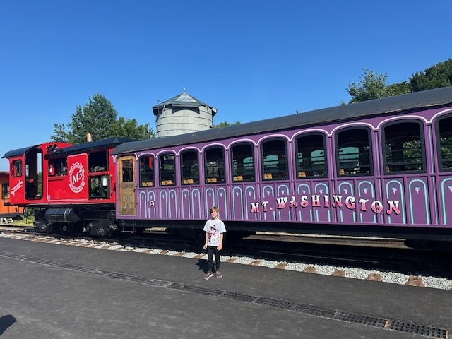 Jensen stands posing in fromt of an old train with a purple car with "Mt. Washington" painted on the side, a red caboose, and a grain tower in the background