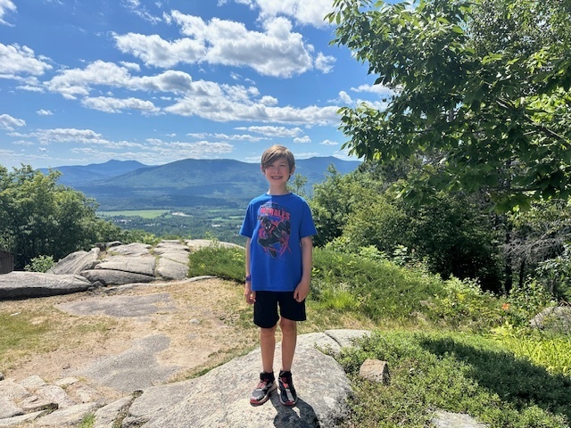 Jensen stands on top of huge rock formations surrounded by trees, overlooking a green valley and mountains