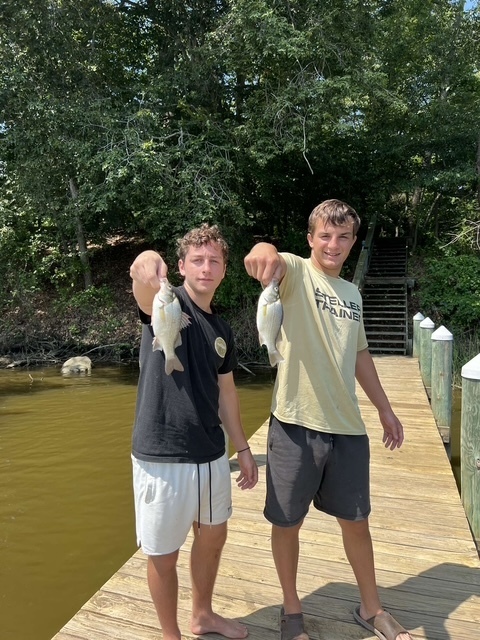 Ryan and Matty hold up the fish they caught while standing on a dock with piers and steps, water on either side, and trees in the background