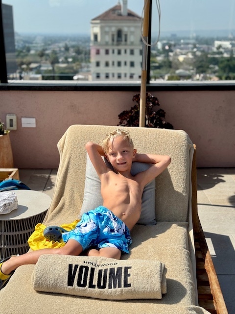 Henrik relaxes on a lawn chair in a blue swimsuit, with buildings in the scenery over a half-wall in the background, and a towel that says "Hollywood Volume" rolled up on the lawn chair in the foreground