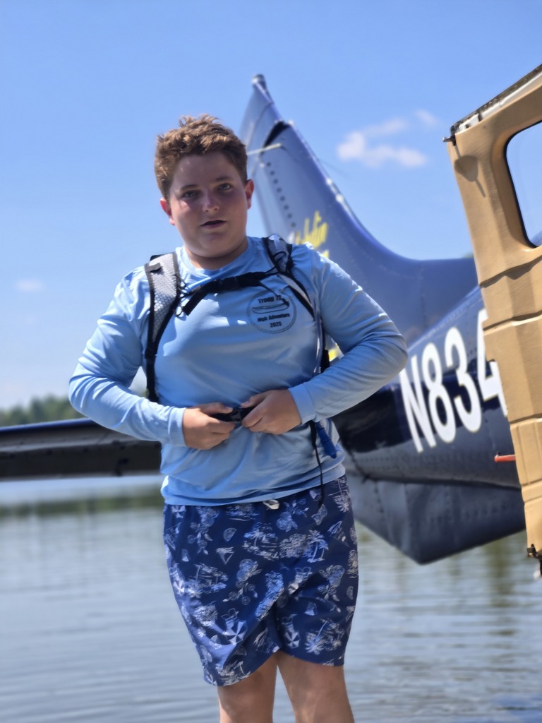 Austin is fastening safety gear around his waist, standing in front of the rear of a sea plane with a body of water beneath
