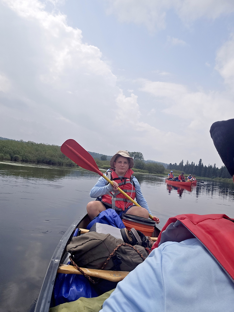 Austin is paddling a canoe filled with duffle bags and gear on a body of water, wearing a hat and safety vest, with another canoe in background