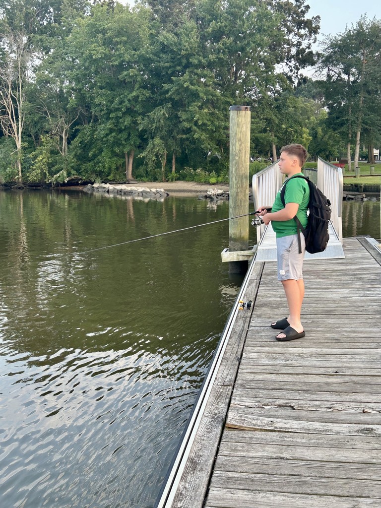 Dominic stands on a dock wearing a green shirt and shorts, fishing over the edge, wearing a black backpack