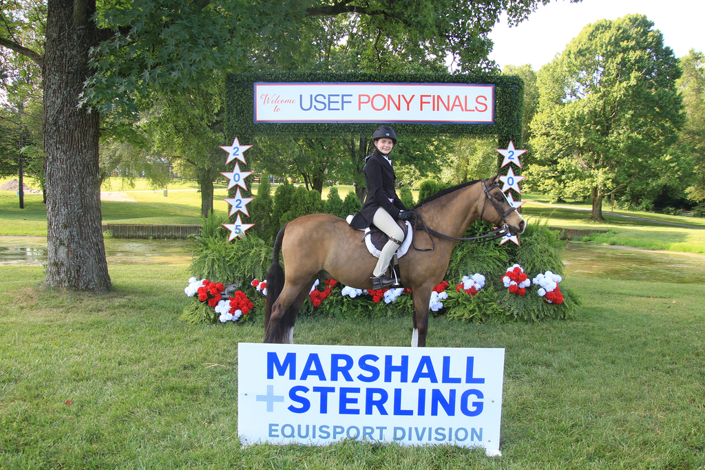 Sophia poses sitting on her pony, in front of a decorated sign saying "Welcome to USEF Pony Finals" and another in the foreground saying "MARSHALL & STERLING EQUISPORT DIVISION"