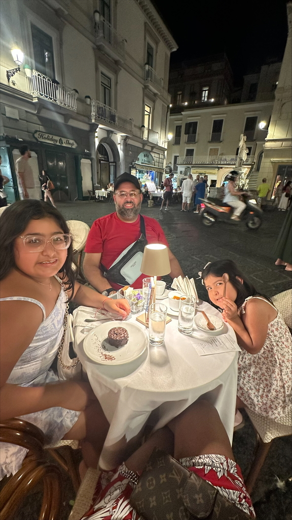 Naomi  sits at a table at an outdoor cafe at nighttime, with her dad and sister, eating dessert