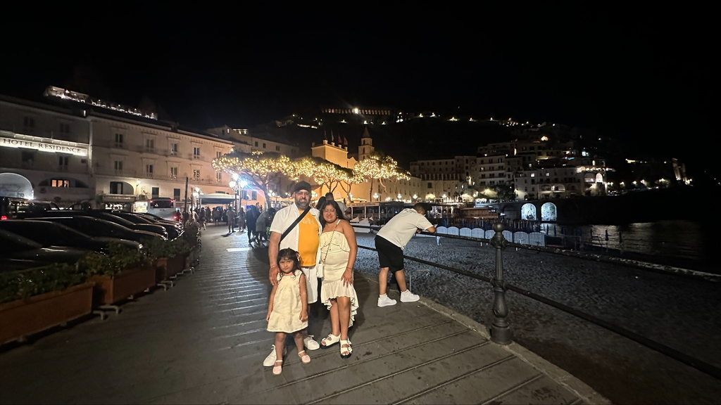 Naomi  with her dad and sister stand on a pier with water beneath, with lights and buildings in the background