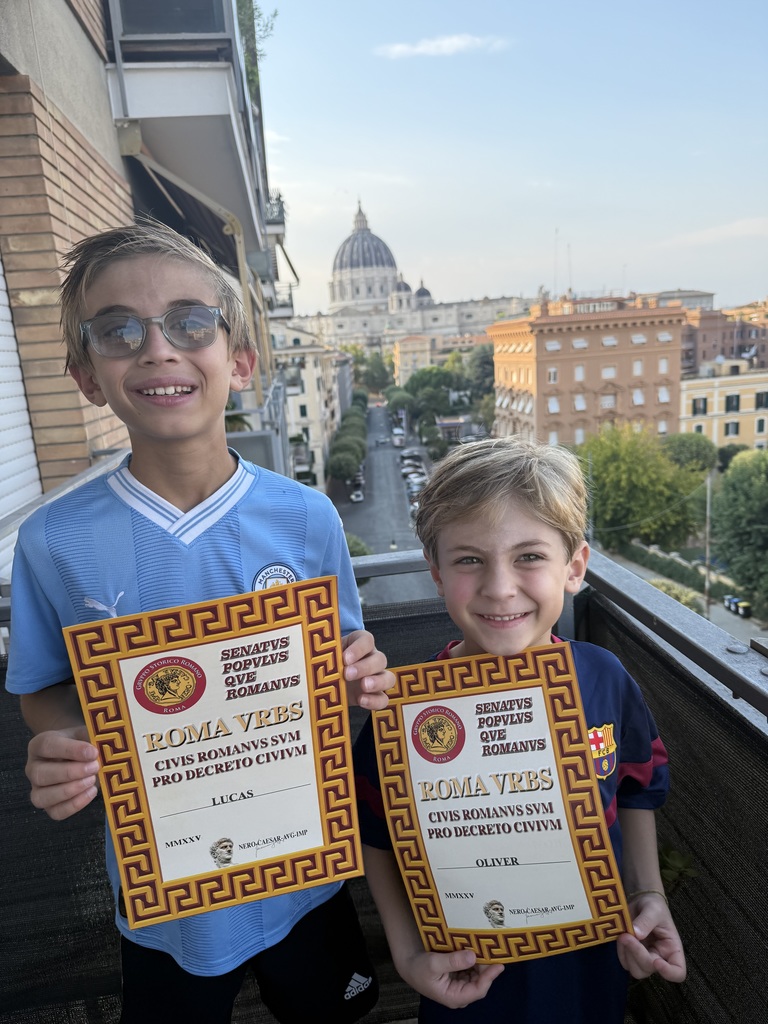 Lucas and Oliver hold up certificates of participation at Gladiator School, standing in a balcony with the Roman city street in the background beneath