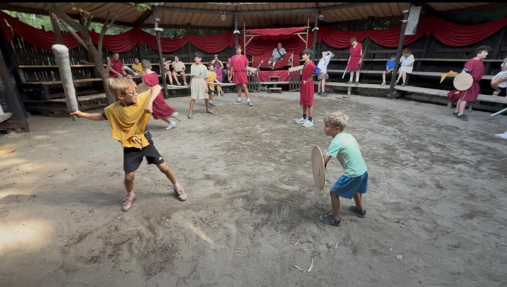 Lucas and Oliver standing in a ring of sand practicing gladiator moves with other young people in the background practicing as well